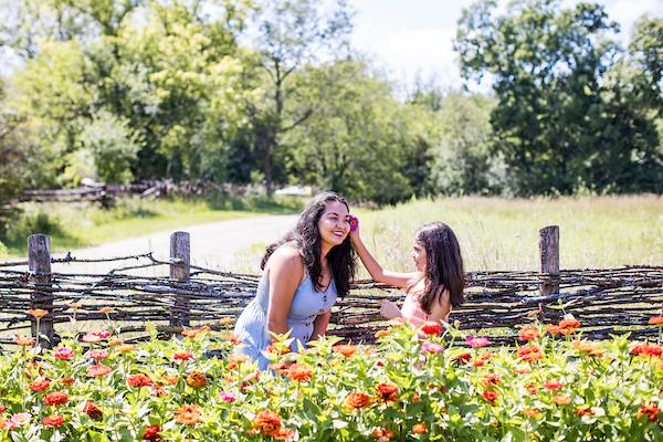 a little girl places a flower in her mother's hair while standing a field of flowers, with a rough woven wooden fence behind them.