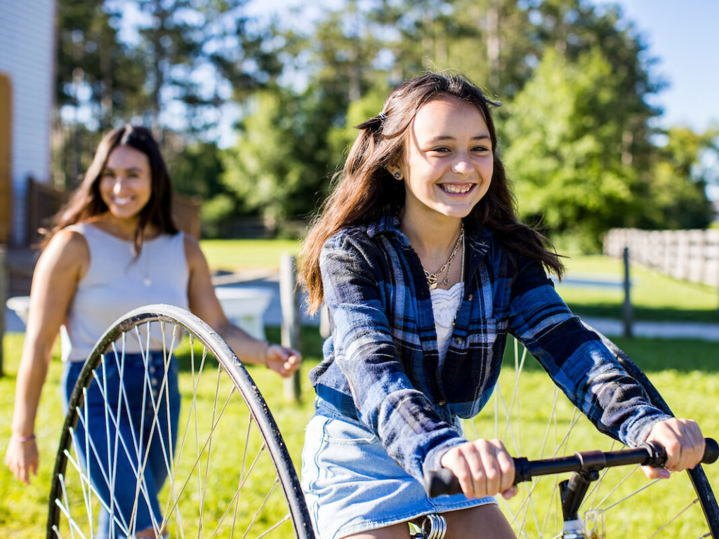A young girl smiles wide as she rides a high wheel tricylce while her equally joyful mom follows behind.