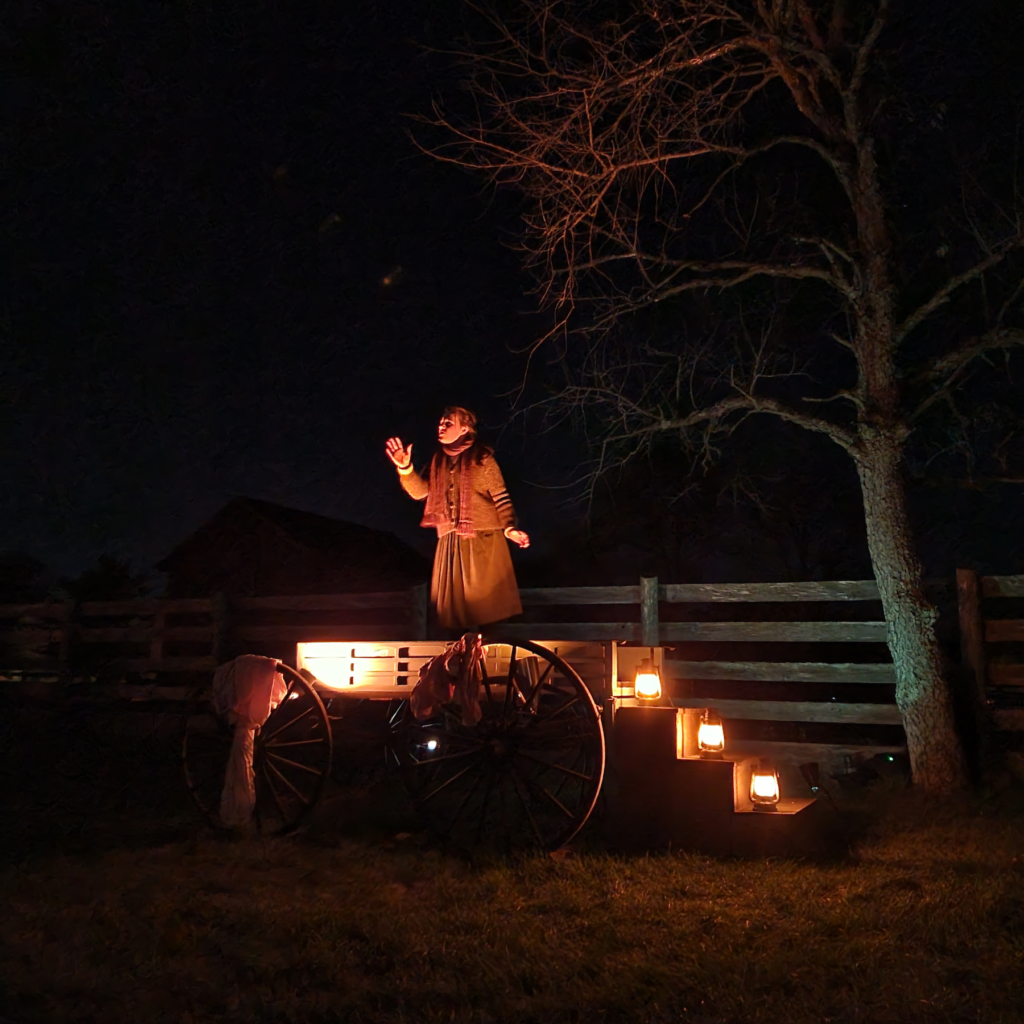 A woman stands on a stage at night, lit from below by orange hued lanterns casting her face in a creepy glow. She is animatedly telling the story of the Beast of Bray Road during the Halloween Legends & Lore event.