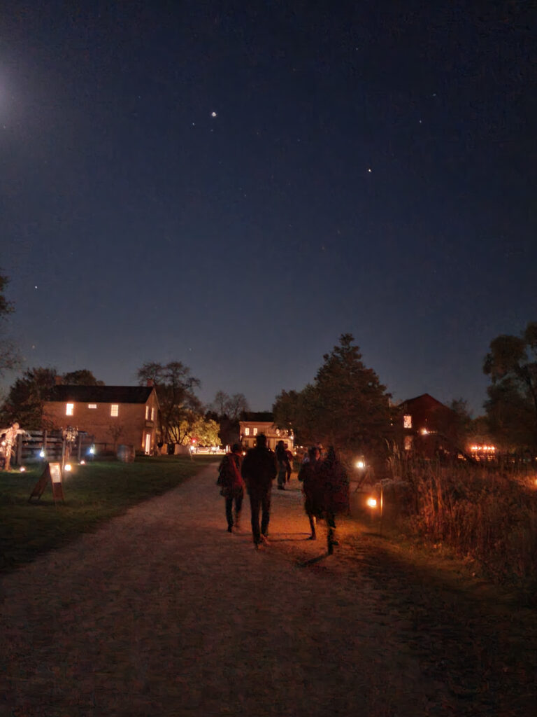 A group of people walk through Cross Roads Village at night with the stars out and the lights casting an eerie glow.